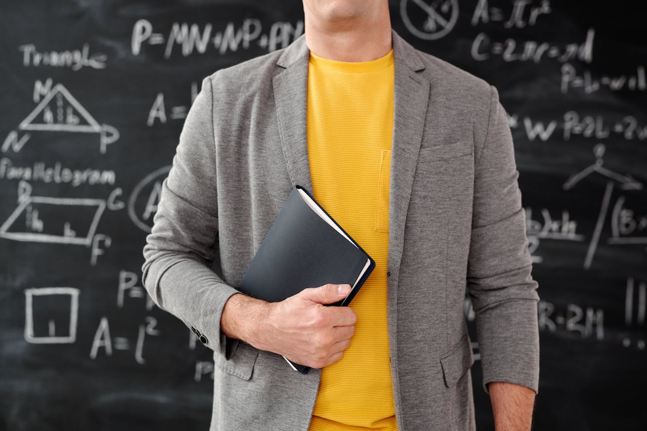 Close-up of a teacher holding a notebook in a classroom setting with equations on the blackboard.
