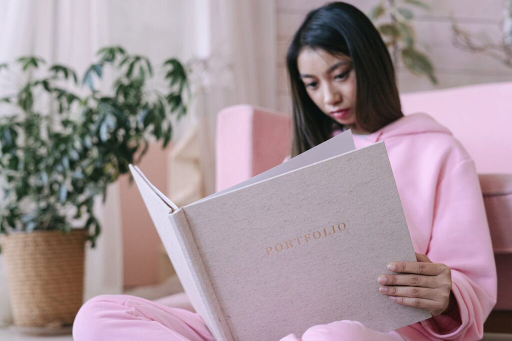 Asian teenager in pink hoodie reading a portfolio indoors with potted plants.